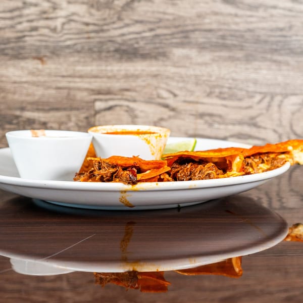 Three birria tacos with two dipping sauces and a lime wedge on a white plate.