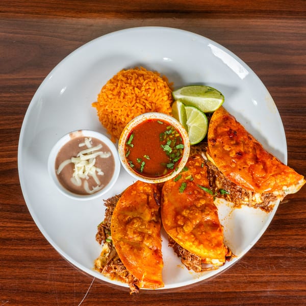 Three birria tacos with rice, refried beans, consome, and lime wedges on a white plate.
