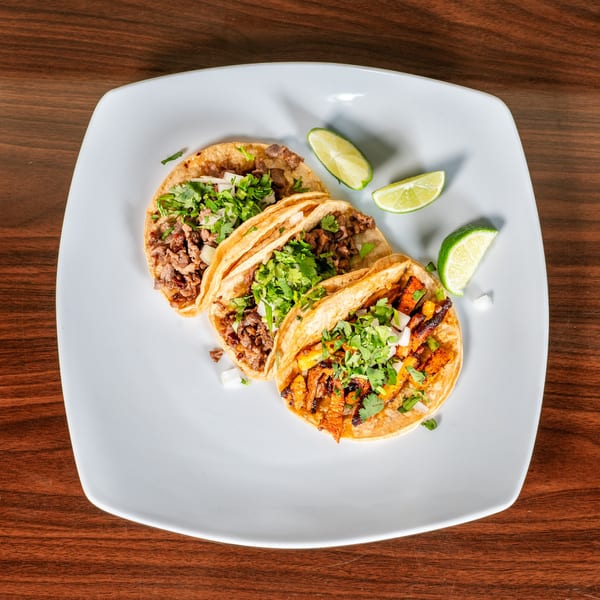 Three delicious Mexican tacos, two with carne asada and one al pastor, garnished with cilantro, onions, and lime wedges.