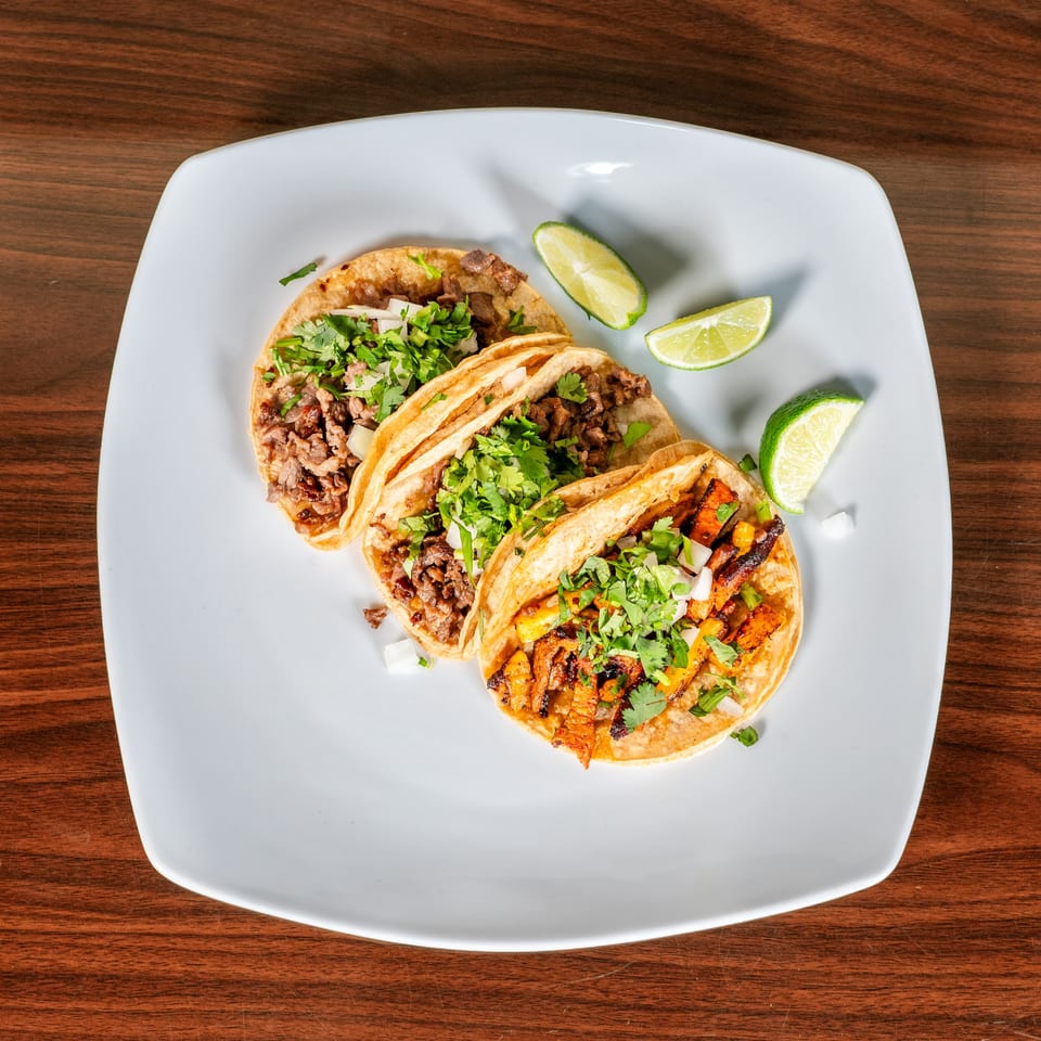 Three delicious Mexican tacos, two with carne asada and one al pastor, garnished with cilantro, onions, and lime wedges.