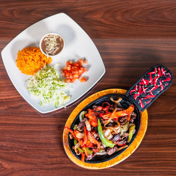 Sizzling beef fajitas with colorful peppers and onions, served with rice, beans, lettuce, and diced tomatoes.