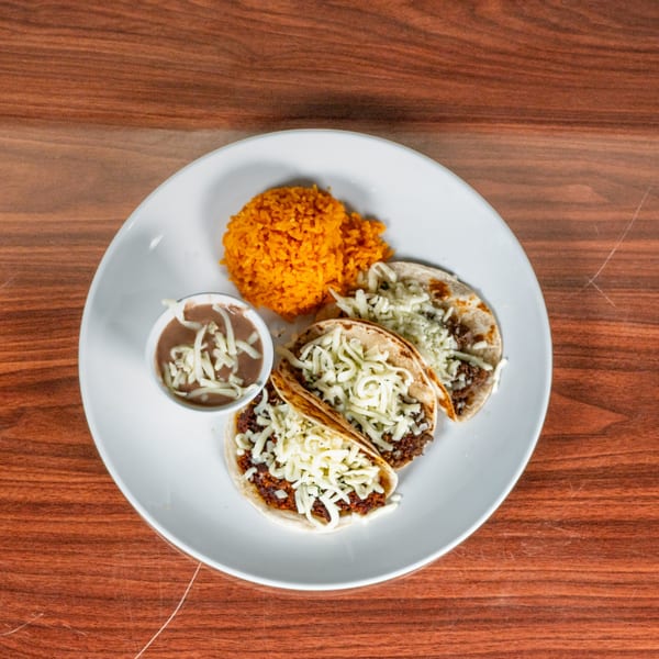 Three tacos with shredded cheese, a side of orange rice, and a small bowl of refried beans.