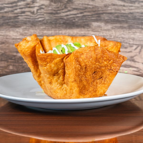 A golden-brown fried taco shell bowl filled with avocado slices and white sauce on a white plate.