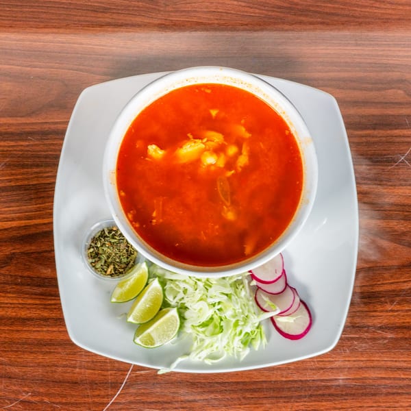 A bowl of red pozole soup with shredded cabbage, lime wedges, radishes, and dried herbs.